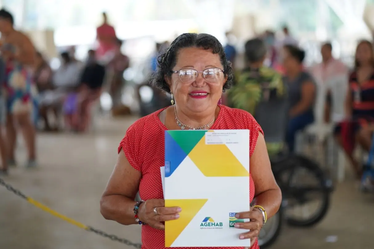 Uma senhora de aproximadamente 60 anos e de blusa vermelha e óculos segurando a escritura concedida pela Agehab.