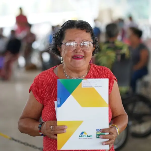 Uma senhora de aproximadamente 60 anos e de blusa vermelha e óculos segurando a escritura concedida pela Agehab.