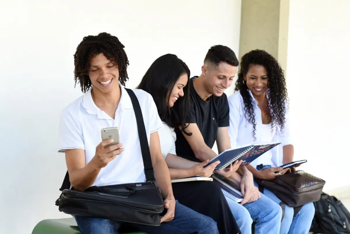 Quatro estudantes, duas moças e dois rapazes com mochilas, livros e celulares, conversando e sorrindo.