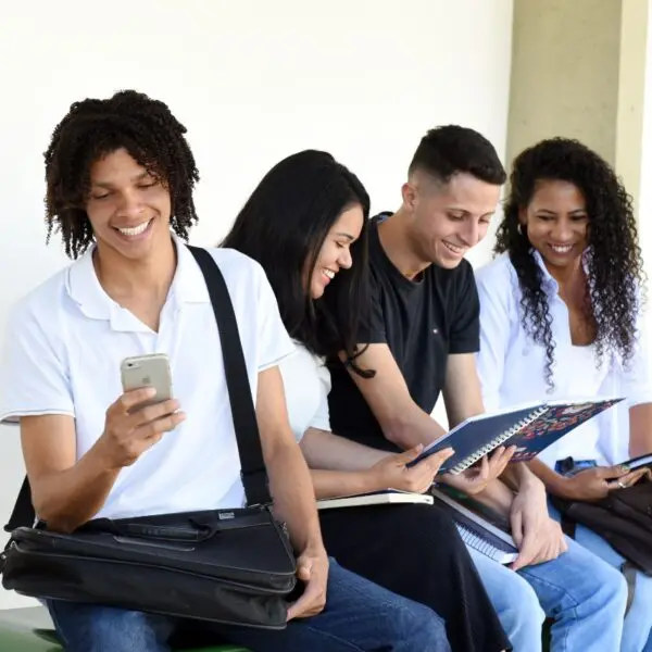 Quatro estudantes, duas moças e dois rapazes com mochilas, livros e celulares, conversando e sorrindo.