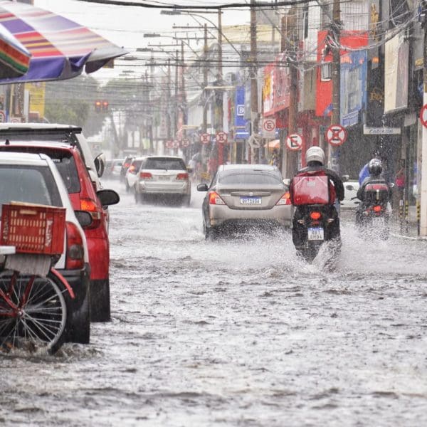 Rua alagada com carros e motos trafegando. Alerta de risco de tempestade